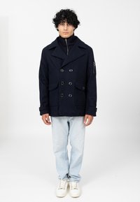 Granatowy wełniany płaszcz typu peacoat z podwójnym zapięciem, dużymi guzikami, kieszeniami z klapką oraz zamkiem na szyi; zestawiony z jasnoniebieskimi dżinsami i białymi trampkami.