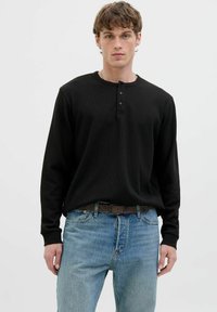 Chemise henley noire à manches longues en tissu texturé, avec trois boutons au col. Associée à un jean en denim bleu clair.