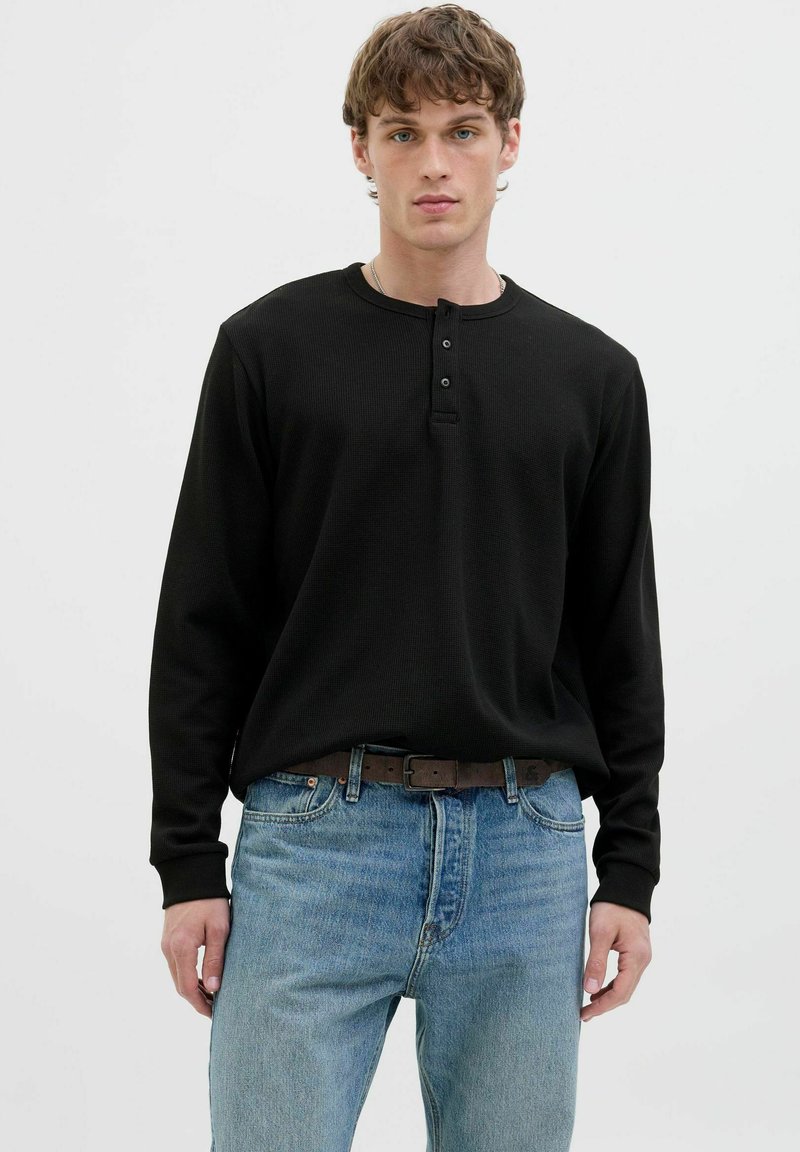 Chemise henley noire à manches longues en tissu texturé, avec trois boutons au col. Associée à un jean en denim bleu clair.