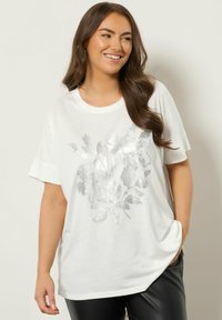 Hvid t-shirt med korte ærmer, prydet med et skinnende sølvfarvet blomstermønster på forsiden. Stoffet ser blødt og let ud.