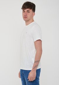 T-shirt bianco a girocollo realizzato in tessuto leggero e testurizzato, con un piccolo logo blu sul lato sinistro del petto. Abbinato a jeans blu.