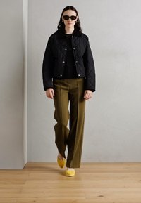 Veste noire matelassée avec des boutons-pression, haut noir, pantalons larges vert olive et chaussures jaunes à enfiler. Les accessoires comprennent des lunettes de soleil noires.