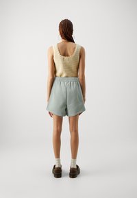 Top in maglia beige con bordi ondulati, abbinato a pantaloni corti verde chiaro e scarpe marroni. Texture lisce e vestibilità comoda.