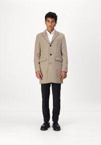 Beige Wollmantel mit tailliertem Design, einreihiger Knopfverschluss, zwei vorderen Taschen und schwarzen Knöpfen, kombiniert mit schwarzen Hosen und Schuhen.