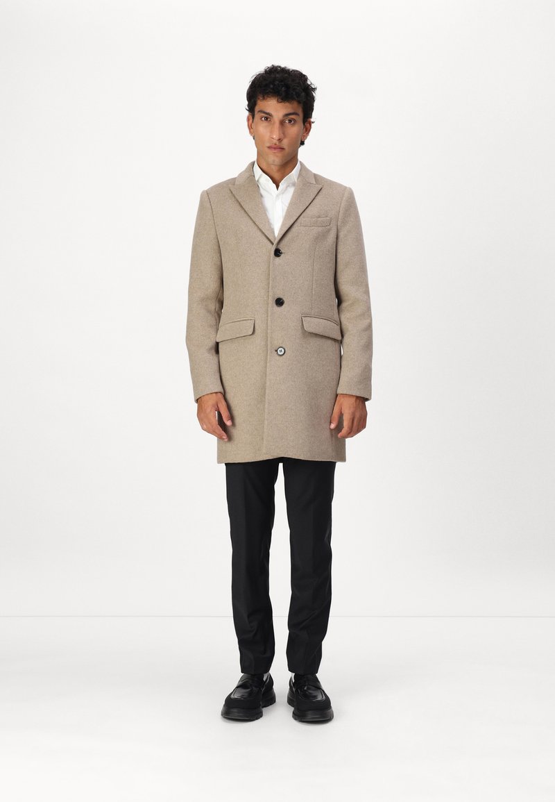 Beige Wollmantel mit tailliertem Design, einreihiger Knopfverschluss, zwei vorderen Taschen und schwarzen Knöpfen, kombiniert mit schwarzen Hosen und Schuhen.