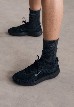 Piernas con zapatillas negras Nike y calcetines negros Nike sobre un suelo beige, un pie ligeramente levantado, textura de piel visible.