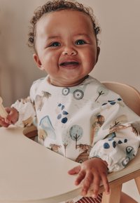 Bavoir à manches longues en tissu blanc orné de motifs multicolores d'animaux et de nature. Enfant assis sur une chaise haute en bois, souriant.