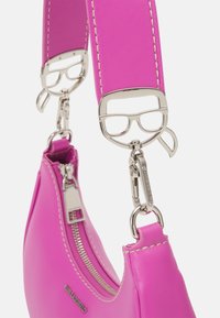 Sac à main en cuir rose avec une forme incurvée, featuring une fermeture éclair en argent et une quincaillerie de sangle distinctive conçue pour ressembler à des lunettes.