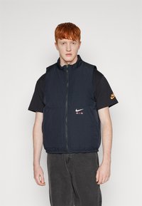 Ναυτική νάιλον αμάνικη γυαλιστερή μπλούζα με ψηλό γιακά, φερμουάρ μπροστά και ελαστικό κάτω μέρος. Διαθέτει λογότυπο Nike και την ένδειξη "AIR" σε πορτοκαλί γράμματα.