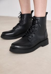 Botas de tobillo de cuero negro con puntera redonda, cierre de cordones en la parte frontal y suela de goma plana. Presenta detalles de costura y una lengüeta en el talón.