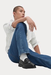 Mocasines de cuero negro con detalle de correa, combinados con jeans de mezclilla azules y calcetines blancos, llevados sobre un fondo claro.