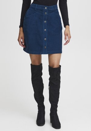 Minikleid aus blauer Cord-Qualität mit vorderem Knopfverschluss, zwei Seitentaschen und einer strukturierten Taille. Kombiniert mit schwarzen Überknie-Stiefeln.