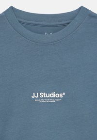 Camiseta azul verdoso hecha de algodón suave. Presenta un cuello redondo y un logo impreso "JJ Studios" en blanco en el pecho. Diseño sencillo y textura lisa.