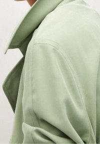 Detailní záběr osoby v oversized světle zeleném trenčkotě s širokým límcem a viditelnými švy na rameni.