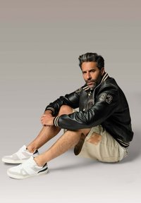 Veste varsity en cuir noir avec col rayé, shorts cargo beiges et baskets blanches avec accents gris. Le sujet est assis contre un fond dégradé doux.