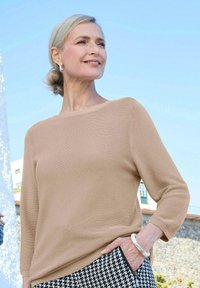 Beiger Strickpullover mit einem Bootsausschnitt und dreiviertellangen Ärmeln, kombiniert mit schwarz-weißen Hahnentritt-Musterhosen.