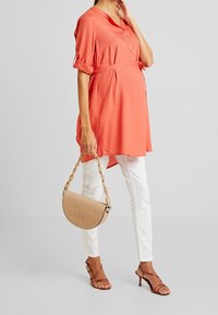 Túnica de color coral con detalles de botones, cintura ceñida con cinturón, combinada con pantalones slim blancos y un bolso de mano trenzado en tono marrón. Sandalias con tiras completan el look.