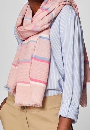 Personne portant une écharpe rayée rose sur une chemise à fines rayures bleu clair et un pantalon beige, les mains dans les poches.