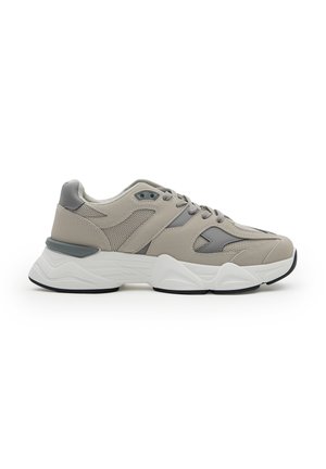 Zapatilla gris y beige con paneles de malla y ante, suela blanca gruesa y cierre con cordones, mostrada de lado sobre un fondo blanco.