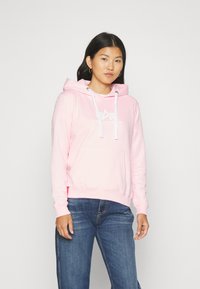 Felpa rosa con tasca frontale, cordini bianchi e stampa del logo. Indossata con jeans blu, per mostrare un look casual e una consistenza morbida.