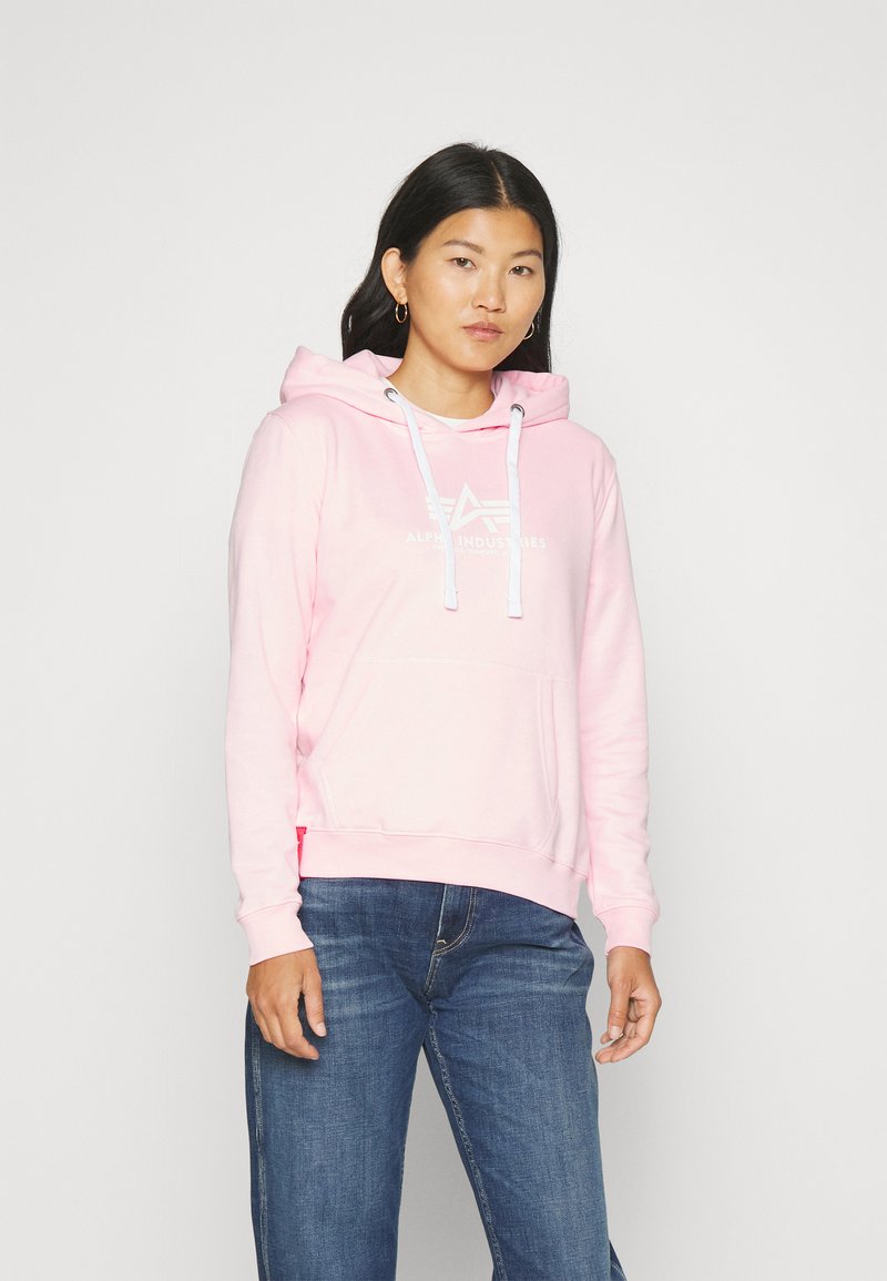 Felpa rosa con tasca frontale, cordini bianchi e stampa del logo. Indossata con jeans blu, per mostrare un look casual e una consistenza morbida.