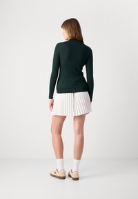 Pull à col roulé côtelé vert foncé, au design ajusté. Assorti à une jupe plissée blanche et des baskets beiges à profil bas avec un détail logo.