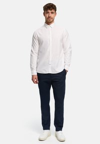 Homme debout, de face, portant une chemise blanche boutonnée, un pantalon bleu marine et des baskets blanches, sur un fond clair uni.