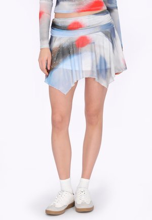 Žena v pestrobarevné asymetrické sukni s červenými, modrými a černými vzory, bílými ponožkami a bílými teniskami stojící na jednotném pozadí.