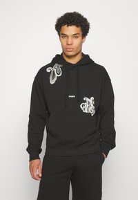 Sweatshirt à capuche noire en coton, avec une poche avant et des motifs paisley colorés sur la poitrine. Logo "HUGO" imprimé à l'avant.