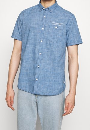 Homme portant une chemise bleue à manches courtes avec boutonnière, poche poitrine et texte blanc, associée à un jean bleu clair, sur fond blanc.