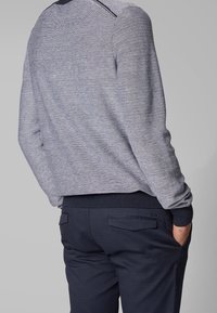 Grå stickad tröja med en texturerad mönster, med marinblå muddar och midjeband, i kombination med mörkblå byxor som visar subtila fickdetaljer.