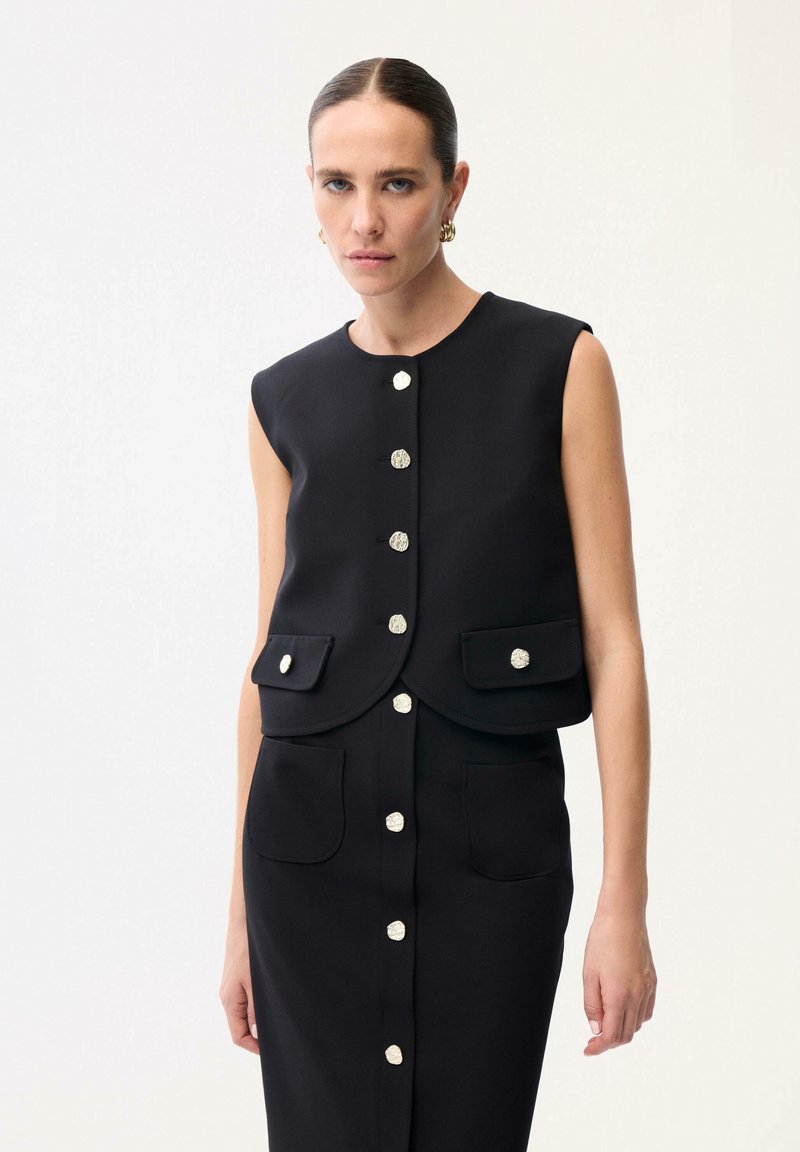 Frau trägt eine ärmellose schwarze Weste mit Knöpfen und passenden Rock mit Vordertaschen vor weißem Hintergrund.