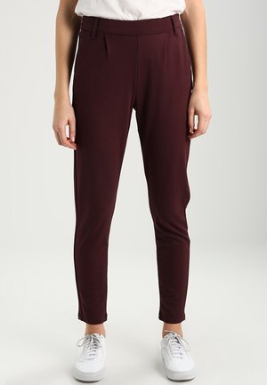 Person wearing fitted dark burgundy trousers and white sneakers, standing against a plain light background.