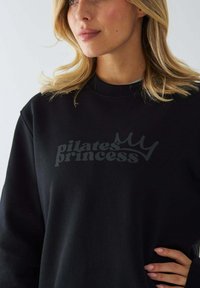 Czarna bluza z matowym napisem "księżniczka pilatesu" z motywem korony. Miękki materiał, okrągły dekolt i długie rękawy.