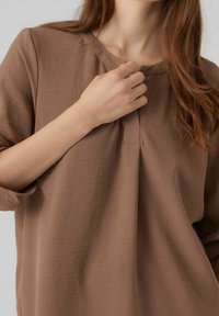 Maglia marrone a maniche lunghe con scollo tondo e pieghe sottili davanti. Tessuto morbido e vestibilità comoda. Dettagli di cuciture visibili.