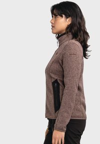 Braune Fleecejacke mit hohem Kragen, seitlichem Reißverschlussfach und kontrastierenden schwarzen Akzenten. Strukturiertes Gewebe mit dezenten Musterdetails.