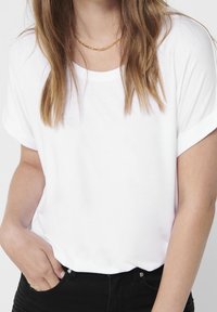 Donna con capelli castano chiaro che indossa una maglietta bianca a maniche corte, pantaloni neri, una collana di catena d'oro e una mano in tasca.