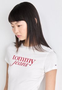 Balta medvilninė marškinėlių su raudonu "tommy jeans" tekstu. Trumpi rankoviai, apvalus iškirpimas ir mažas logotipas ant rankovės. Paprastas dizainas.