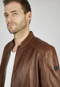 Chaqueta de cuero marrón con cuello alzado, cierre de cremallera y acentos acolchados en los hombros. Presenta un logo redondo negro en la manga izquierda.