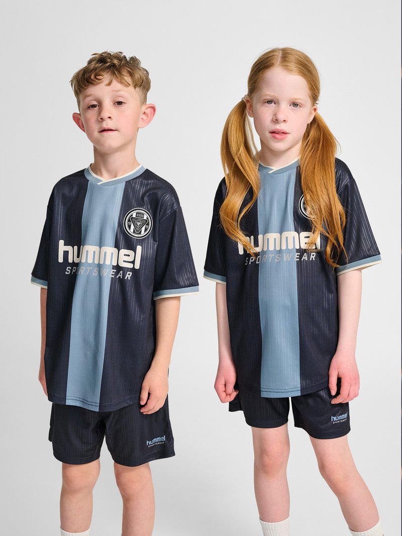 Twee kinderen dragen zwarte en blauwe sportkleding met het "hummel"-logo, staand tegen een effen grijze achtergrond.