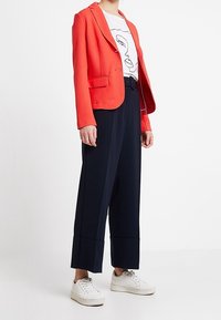 Blazer en coral avec un tissu texturé, un bouton unique et deux poches avant, associé à un t-shirt graphique blanc, un pantalon large bleu marine et des baskets blanches.