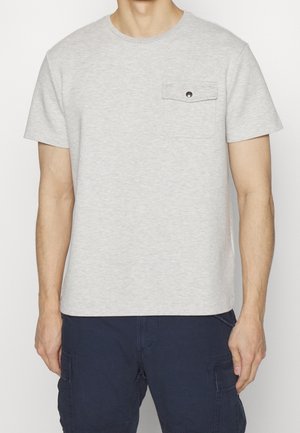 Homme portant un T-shirt à manches courtes gris clair avec une poche poitrine boutonnée et un pantalon cargo bleu foncé, debout devant un fond clair uni.