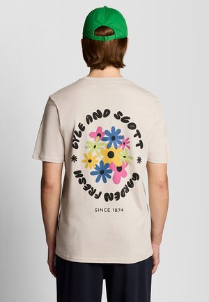 Person trägt ein beigefarbenes T-Shirt mit bunten Blumenmotiven und dem Text „Cyle and Scott Garden Fresh Since 1874“ auf der Rückseite sowie eine grüne Mütze.