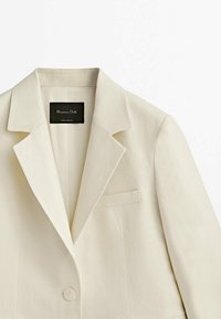 Cremefarbener Blazer mit Reverskragen, Einzelknopfverschluss und Brusttasche. Hergestellt aus strukturiertem Stoff. Etikett innen sichtbar.