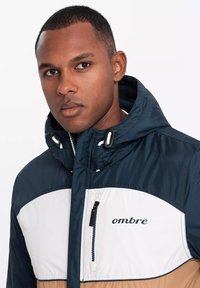 Marineblau, weiß und beige farblich abgesetzte Jacke mit Kapuze, ausgestattet mit einer mit Reißverschluss versehenen Brusttasche und einem Logodetail auf der Vorderseite. Glattes, glänzendes Material.