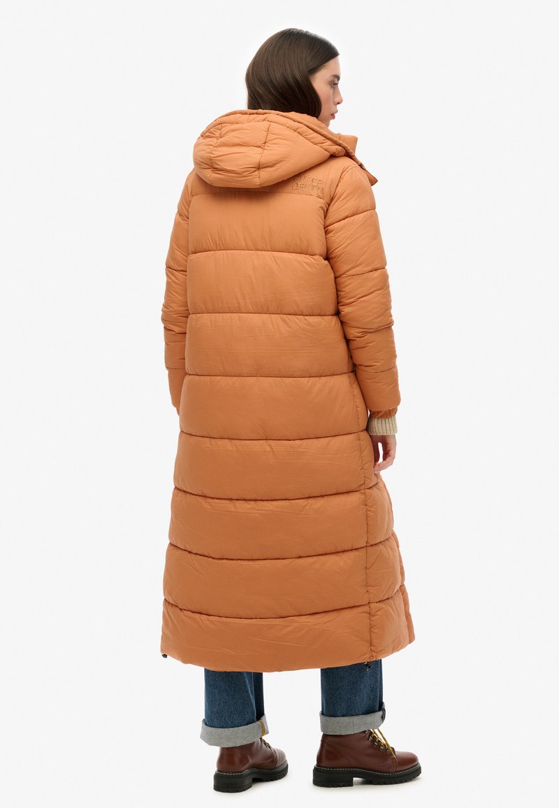 Lange pufferjas in warm oranje met horizontaal gestikt ontwerp, verstelbare capuchon en zijzakken, gecombineerd met blauwe jeans en bruine laarzen.