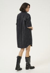 Vestido de denim negro oversize con mangas cortas y dobladillo sin terminar, combinado con botas de cuero negro hasta la rodilla con hebillas.