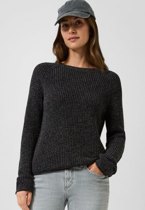 Femme portant un pull en maille gris foncé, un jean gris clair et une casquette de baseball grise, une main dans la poche.