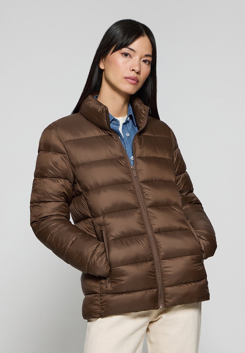 Brązowa kurtka puchowa z wysokim kołnierzem, zapięciem na zamek, poziomym pikowaniem i bocznymi kieszeniami. Nosi na niebieskiej koszuli. Gładka tekstura.