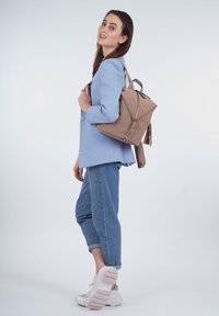 Mochila de tela beige con diseño triangular y herrajes plateados, que presenta una cremallera y acentos de borlas, llevada de manera casual con un blazer azul y jeans.